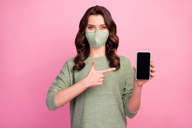 Portrait d'une fille gaie positive, promoteur de point d'index, smartphone portant un masque médical cavalier isolé sur fond de couleur pastel