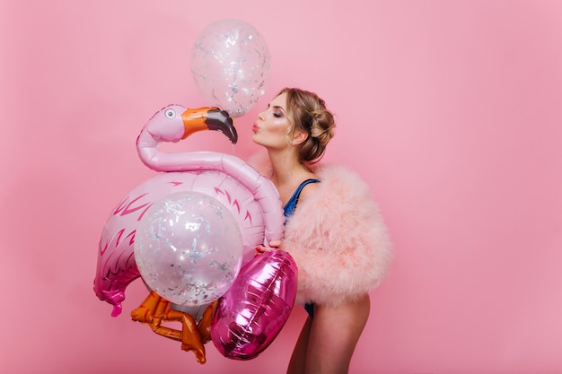 Portrait De Fille Gracieuse Mince Aux Cheveux Blonds Courts Embrassant Flamant Rose Gonflable. Superbe Jeune Femme Gaie En Manteau Moelleux S'amusant Avec Des Ballons Scintillants, En Attente De Fête