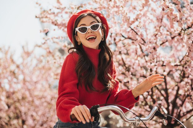 Portrait de fille magnifique en tenue rouge et lunettes de soleil sur fond de sakura. Femme joyeuse en pull en cachemire et béret souriant et vélo