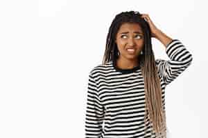 Photo gratuite portrait d'une fille noire confuse qui a l'air maladroite et incertaine en se grattant la tête et en regardant de côté avec une expression de visage indécise sur fond blanc