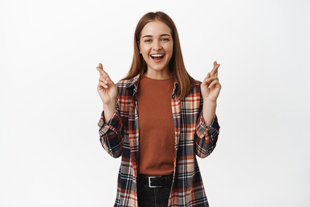 Le portrait d'une fille pleine d'espoir lui fait souhaiter, a l'air heureuse et souriante, croise les doigts pour la bonne chance, prie, anticipe les nouvelles positives, debout sur fond blanc.