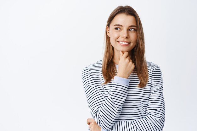 Portrait d'une fille réfléchie faisant l'hypothèse, ayant des idées intéressantes, regardant à gauche le logo de l'espace de copie et souriant, méditant, debout sur un mur blanc