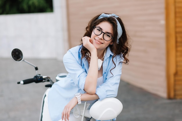 Portrait d'une fille rêveuse dans des verres avec des boucles sombres brillantes soutenant le visage avec la main, se relaxant sur un scooter. Jolie jeune femme en montre-bracelet tendance à la recherche avec intérêt et sourire mignon.