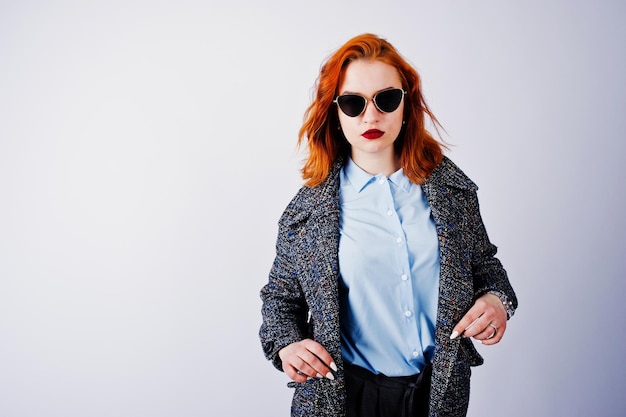 Portrait d'une fille rousse fantastique en chemise bleue pardessus gris posant avec des lunettes de soleil dans le studio