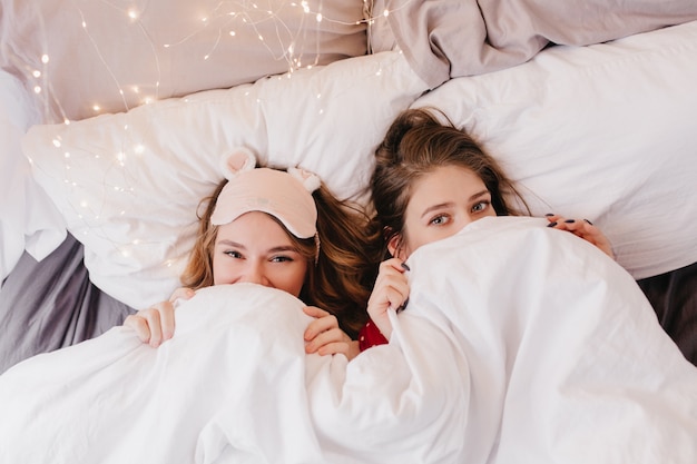 Portrait de frais généraux de jeunes femmes excitées au lit. Sœurs rêveuses se cachant sous une couverture tôt le matin.