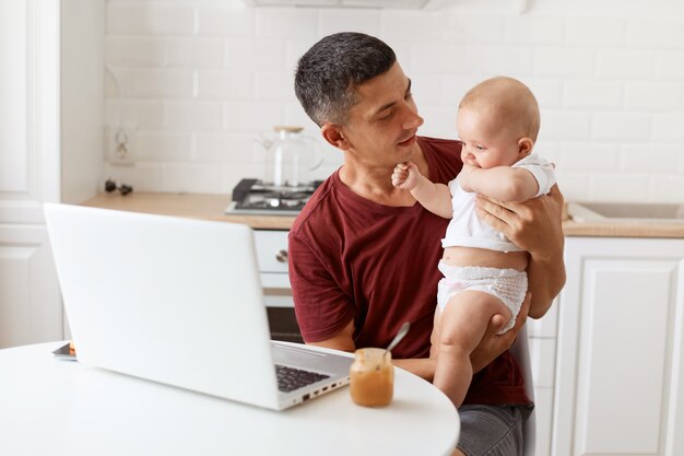 Portrait d'un freelancer séduisant homme brune portant un t-shirt marron de style décontracté, travaillant et s'occupant de sa petite fille, regardant bébé avec amour.