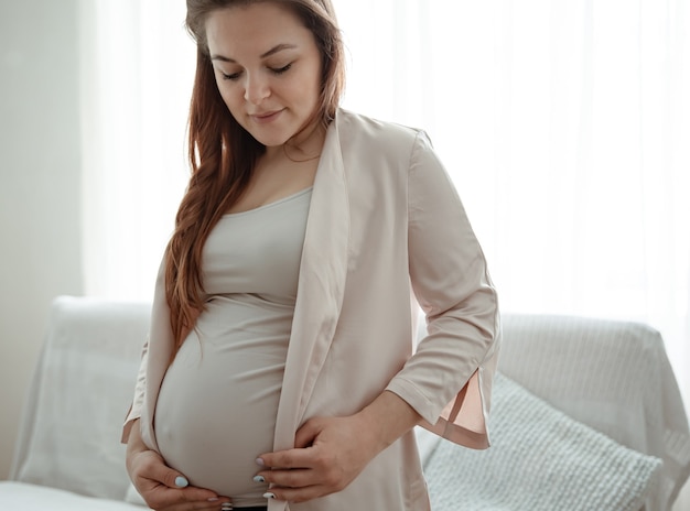 Portrait d'une future maman avec un gros ventre dans le salon près de la fenêtre.