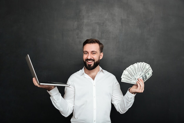 Portrait de gai homme riche en chemise blanche gagnant beaucoup d'argent monnaie dollar à l'aide de son carnet sur gris foncé