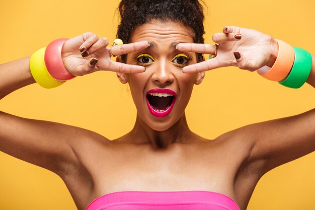 Photo gratuite portrait gai de jeune femme mulâtre souriante avec maquillage lumineux montrant signe de paix avec les doigts sur l'appareil photo, isolé sur mur jaune