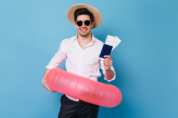 Portrait de gars au chapeau et lunettes sur l'espace bleu. Homme d'affaires en chemise détient des billets pour le repos, le passeport et l'anneau en caoutchouc.