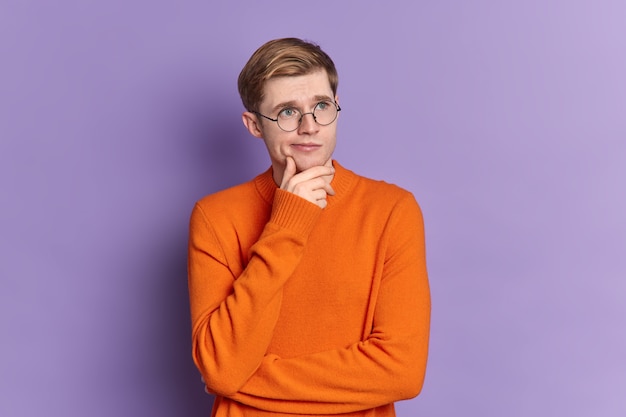 Portrait de gars aux yeux bleus réfléchit à quelque chose se dresse dans une pose réfléchie tient le menton concentré dans la distance porte un cavalier orange occasionnel