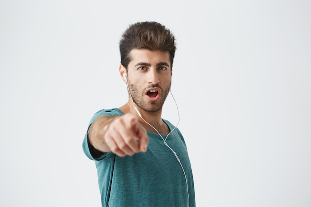 Portrait d'un gars barbu élégant ayant une coiffure à la mode portant un T-shirt bleu et des écouteurs blancs avec la bouche ouverte et pointant vers vous. Concept de personnes et d'émotions