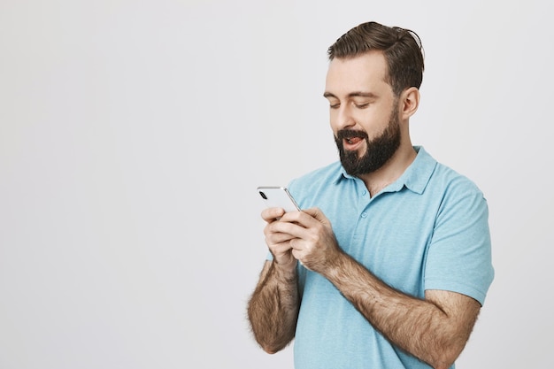 Portrait d'un gars drôle avec une barbe en regardant son téléphone debout sur blanc. l'homme essaie d'inventer un message amusant pour féliciter son meilleur ami avec son anniversaire.