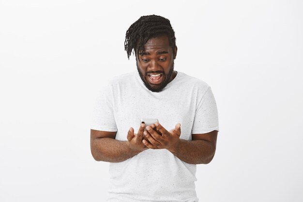 Portrait de gars excité avec des tresses posant contre le mur blanc avec son téléphone