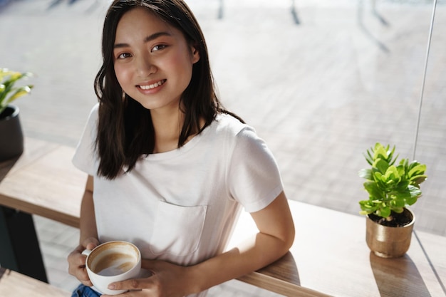 Portrait en grand angle d'une fille asiatique souriante, buvant du café dans un café près de la fenêtre, regardant heureux tenant une tasse de cappuccino