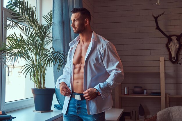 Portrait d'un grand homme barbu au corps musclé, vêtu d'une chemise déboutonnée et d'un jean, se prépare pour une journée de travail.