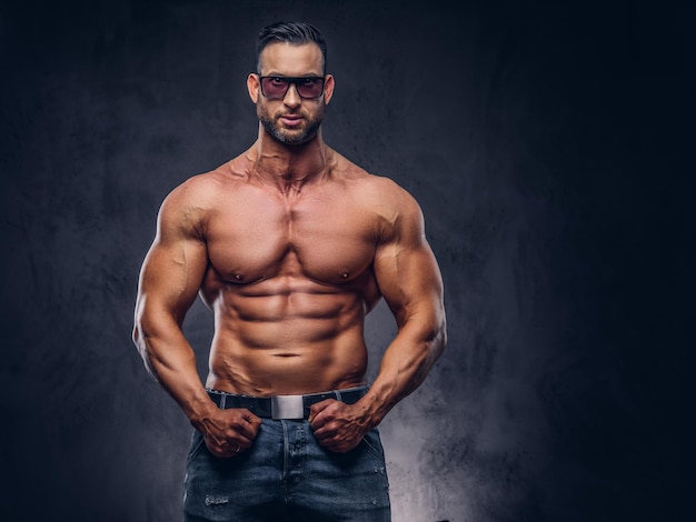 Portrait d'un grand mâle torse nu avec un corps musclé avec une coupe de cheveux et une barbe élégantes, dans des lunettes de soleil et un jean, posant dans un studio. Isolé sur un fond sombre.