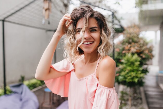 Portrait de gros plan d'adorable jeune femme posant avec un sourire surpris dans un restaurant de rue. Fille bronzée en chemisier rose jouant avec ses cheveux ondulés légers.