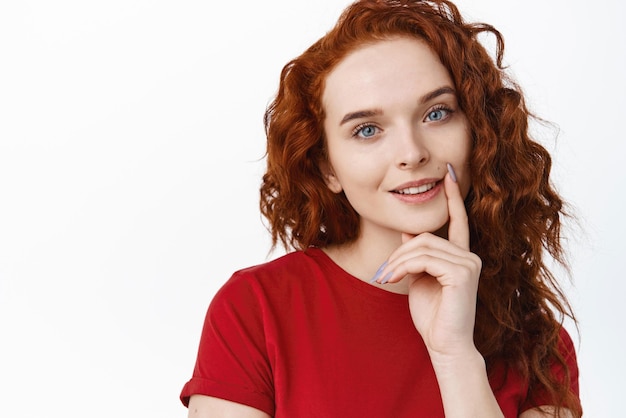 Photo gratuite portrait en gros plan d'un beau modèle féminin rousse avec une longue coiffure bouclée touchant la lèvre et un sourire affectueux à la caméra, l'air pensif et intrigué, debout sur fond blanc