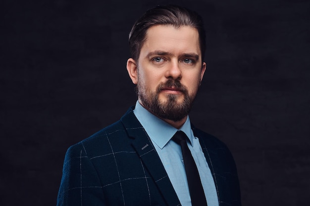 Portrait en gros plan d'un bel homme d'âge moyen avec barbe et coiffure vêtu d'un élégant costume formel sur un fond sombre texturé en studio.