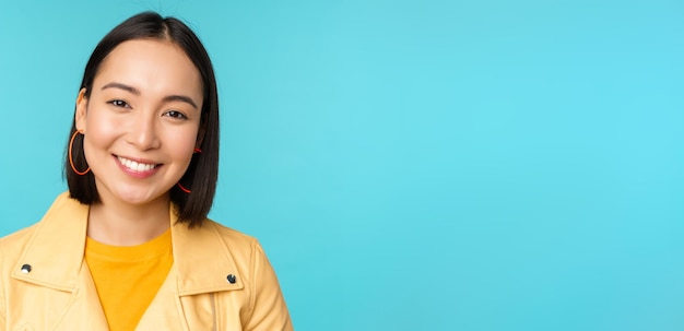 Photo gratuite portrait en gros plan d'une belle femme asiatique souriante aux dents blanches qui a l'air heureuse devant la caméra posant en veste jaune sur fond de studio bleu