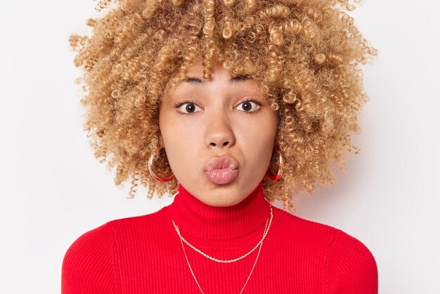 Portrait en gros plan d'une belle femme aux cheveux bouclés blonds garde les lèvres pliées veut vous embrasser a un regard affectueux sur la caméra exprime l'amour porte un col roulé rouge isolé sur fond blanc