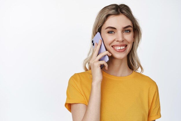 Portrait en gros plan d'une belle femme caucasienne ayant un appel téléphonique tenant un smartphone et souriant à la caméra debout en t-shirt jaune sur fond blanc