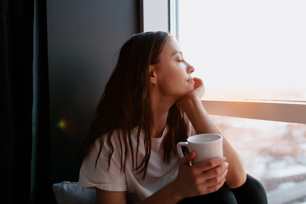 Portrait en gros plan d'une belle femme romantique aux cheveux noirs avec des cheveux ondulés est les yeux fermés et souriant tout en buvant du café le matin et assis près de la fenêtre au soleil du matin