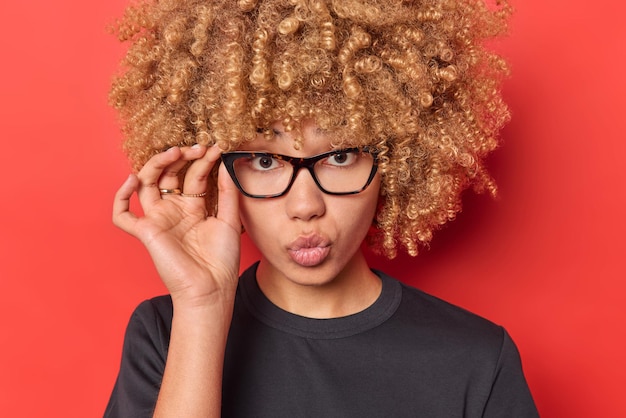 Portrait en gros plan d'une belle jeune femme aux cheveux bouclés garde la main sur le bord des lunettes garde les lèvres pliées regarde attentivement la caméra vêtue d'un t-shirt noir décontracté isolé sur fond rouge