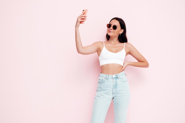 Portrait en gros plan d'une belle jeune femme souriante dans des vêtements d'été à la mode Sexy femme insouciante posant près d'un mur rose en studio Modèle positif s'amusant à l'intérieur Prendre des photos de selfie Dans des lunettes de soleil