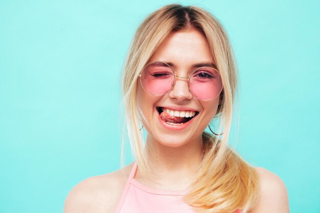 Portrait en gros plan d'une belle jeune femme souriante dans des vêtements hipster d'été à la mode Sexy femme insouciante posant sur fond bleu en studio Modèle positif fait un clin d'œil et montre la langue