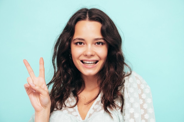 Portrait en gros plan d'une belle jeune femme souriante en robe d'été à la mode Sexy femme insouciante posant près d'un mur bleu en studio Modèle positif s'amusant à l'intérieur Montre un signe de paix