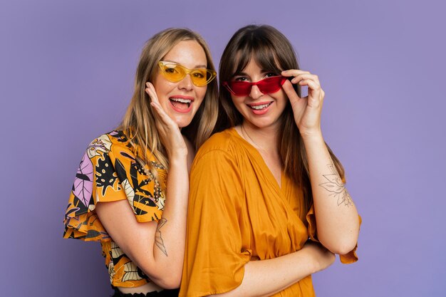 Portrait en gros plan de deux femmes élégantes en lunettes de soleil et vêtements d'été à la mode posant sur fond violet en studio