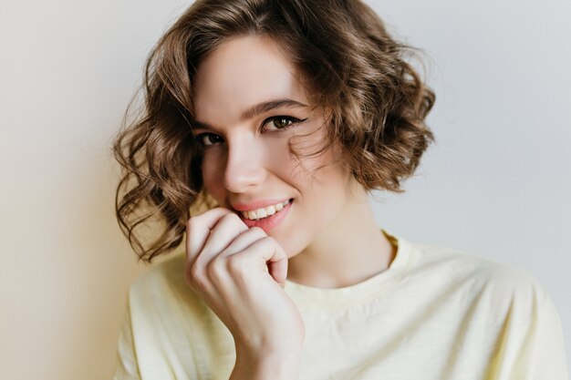 Portrait en gros plan du magnifique modèle féminin avec de beaux yeux. Incroyable fille brune en chemise blanche posant avec le sourire sur un mur lumineux.