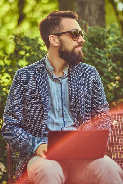 Portrait en gros plan d'un écrivain avec une barbe pleine et une coupe de cheveux élégante, travaillant avec un ordinateur portable tout en étant assis sur un banc dans un parc.