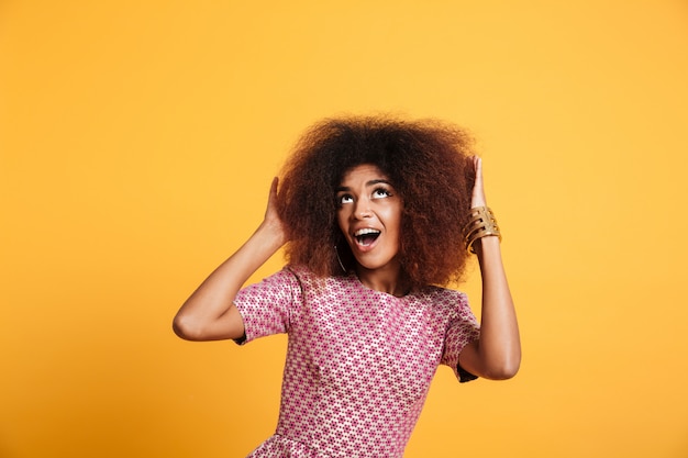 Portrait de gros plan d'une femme africaine étonnée en robe touchant sa coiffure afro, regardant vers le haut