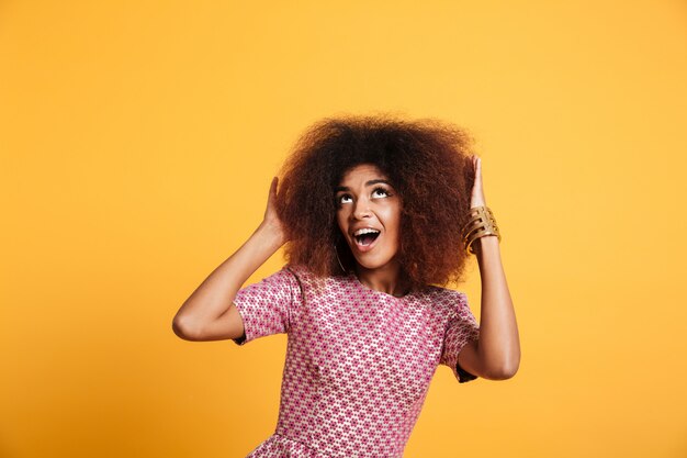 Portrait de gros plan d'une femme africaine étonnée en robe touchant sa coiffure afro, regardant vers le haut