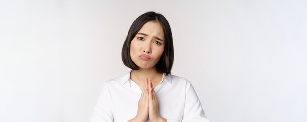 Portrait en gros plan d'une femme asiatique mendiant demandant smth dire s'il vous plaît avec un visage triste timide besoin d'aide debout sur fond blanc