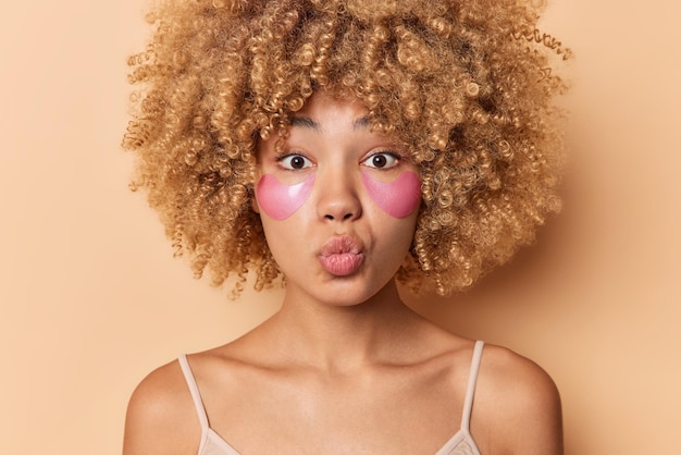 Portrait en gros plan d'une femme aux cheveux bouclés garde les lèvres pliées veut embrasser quelqu'un applique des patchs d'hydrogel rose sous les yeux pour le rajeunissement porte un t-shirt décontracté isolé sur fond beige.