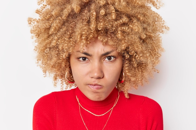 Portrait en gros plan d'une femme aux cheveux bouclés mécontente, les lèvres des sacs à main regarde avec colère la caméra porte un col roulé rouge se sent indigné ou agacé exprime des émotions négatives isolées sur fond blanc.