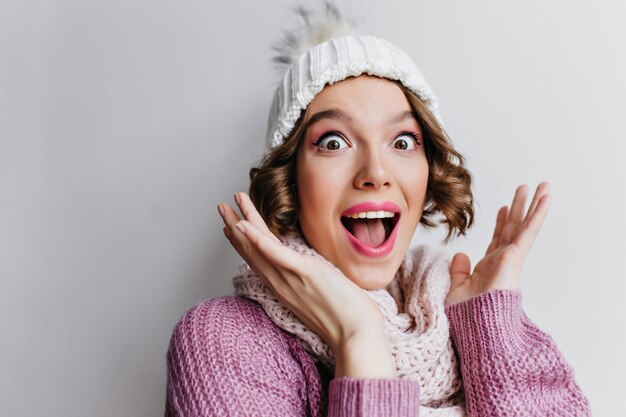 Portrait de gros plan d'une femme aux cheveux courts étonnée avec des lèvres roses posant en chapeau d'hiver. fille blanche surprise en pull violet.