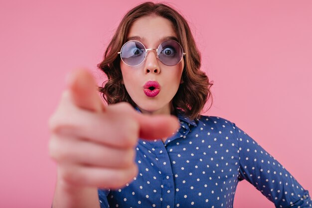 Portrait en gros plan d'une femme aux cheveux courts étonnée porte des lunettes bleues. Jeune femme en chemise élégante exprimant des émotions surprises sur le mur rose.