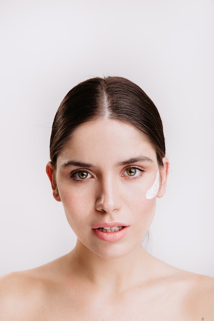 Portrait de gros plan d'une femme aux cheveux noirs aux yeux verts avec une peau saine et de la crème sur son visage. Fille sans maquillage sur un mur blanc.