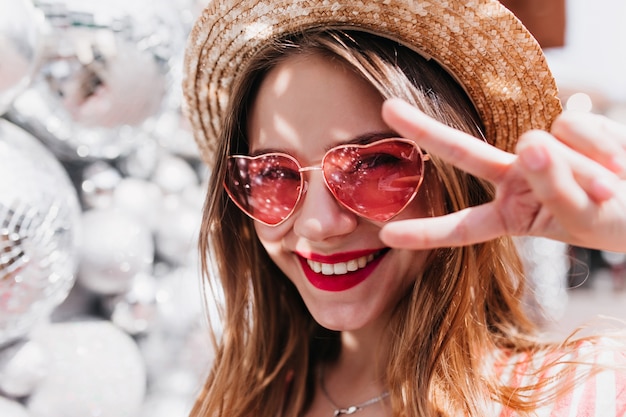 Portrait de gros plan de femme blanche de bonne humeur posant avec signe de paix. Photo de belle fille détendue porte un chapeau et des lunettes de soleil roses.