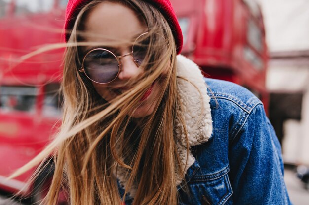 Portrait en gros plan d'une femme blanche séduisante s'amusant dans une bonne journée de printemps. Enchanteur fille caucasienne en veste en jean élégante riant près de bus rouge.