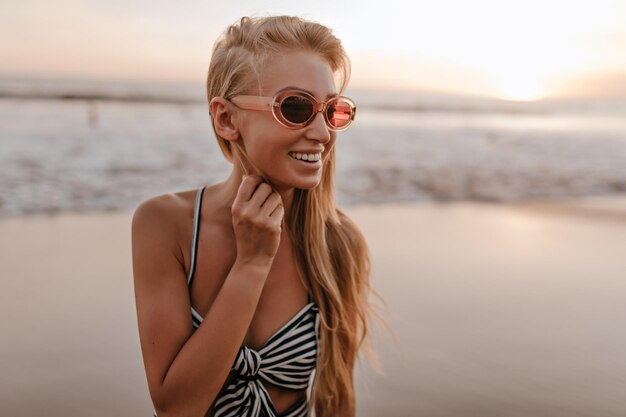 Portrait En Gros Plan D'une Femme Blonde Bronzée Portant Des Lunettes De Soleil Roses Et Un Haut Court à Rayures Sourit Sincèrement Et Profite Du Coucher De Soleil Sur La Plage