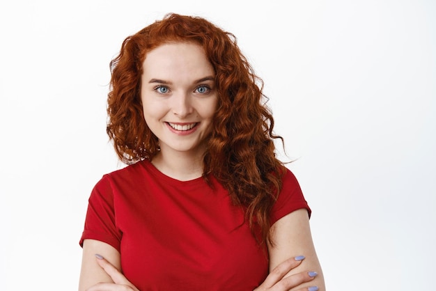 Photo gratuite portrait en gros plan d'une femme confiante aux cheveux bouclés au gingembre, bras croisés sur la poitrine et souriante, peau pâle sans imperfections et yeux bleus sur fond blanc