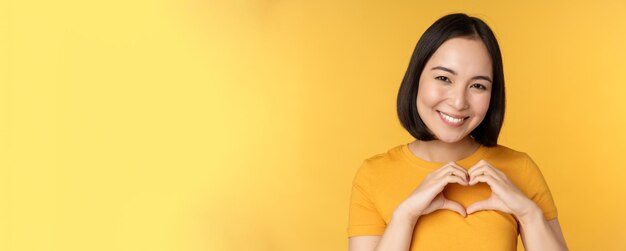 Portrait en gros plan d'une femme coréenne souriante montrant un signe de coeur romantique et ayant l'air heureux debout sur fond jaune