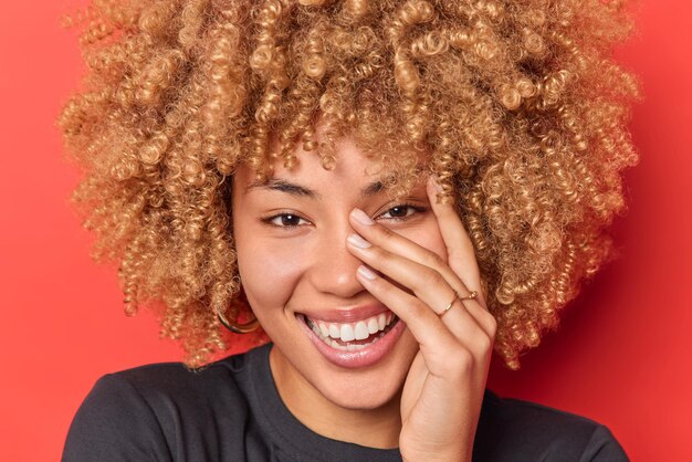 Portrait en gros plan d'une femme heureuse aux cheveux bouclés qui garde la main sur les sourires du visage exprime largement des sentiments sincères avec des yeux pleins de joie isolés sur fond rouge. Notion d'émotions positives