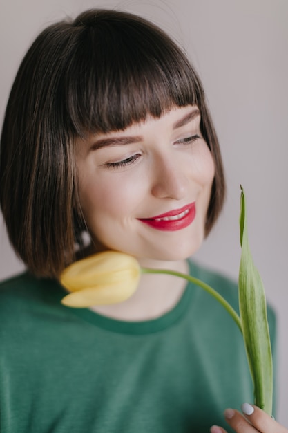 Portrait de gros plan d'une femme heureuse avec des lèvres rouges rêveuses en détournant les yeux tout en posant avec une fleur. Belle fille blanche tenant une tulipe jaune avec un sourire inspiré.